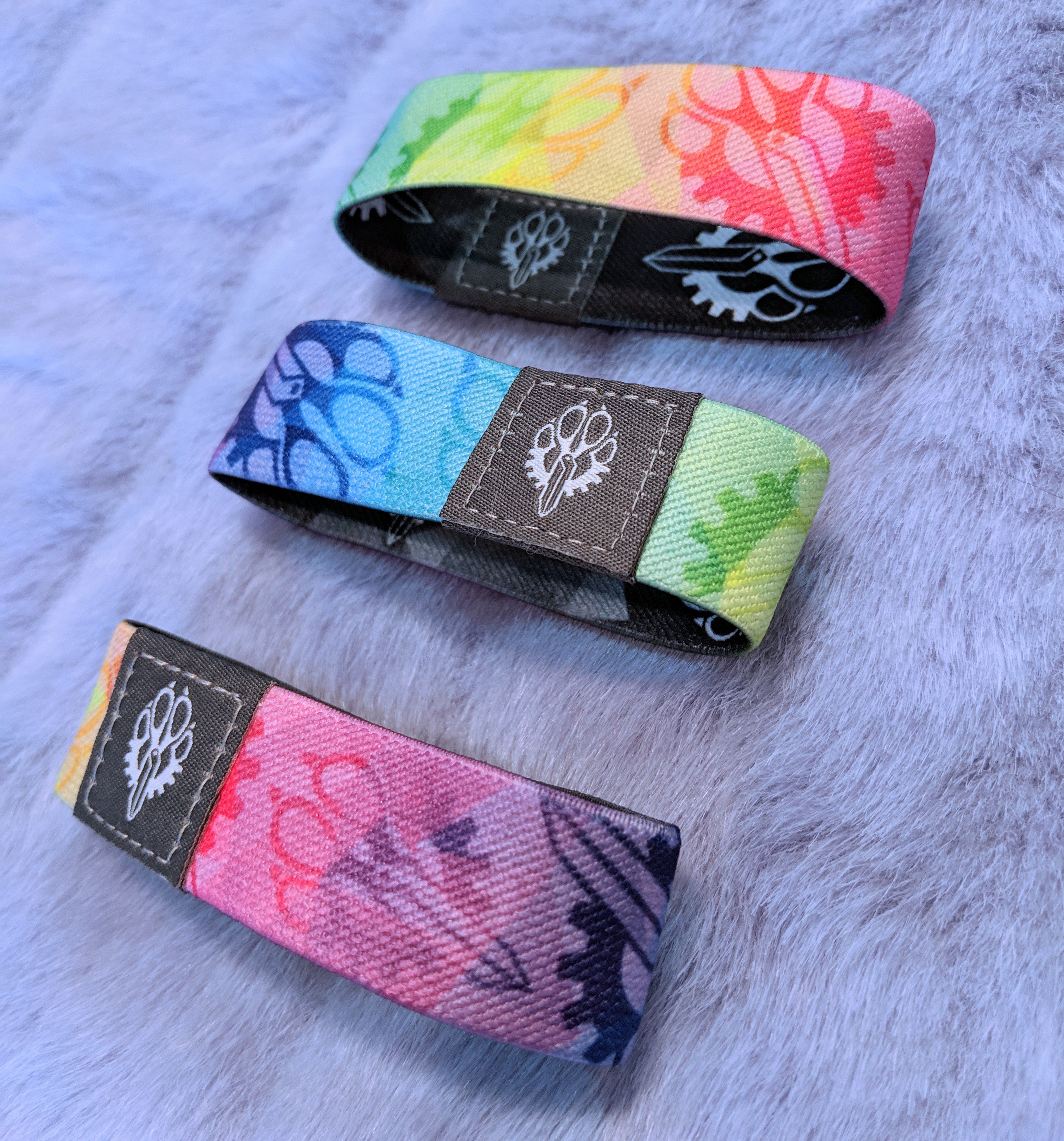 Four colorful Fuzz Factory wristbands with geometric rainbow patterns on a fur surface